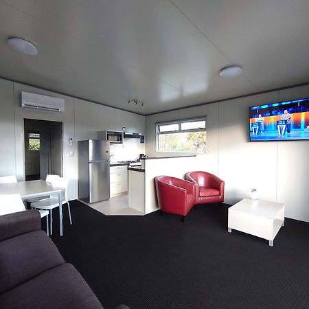 Taupo Debretts Spa Resort Luaran gambar The photo displays a modern, compact living space. It features a small kitchen area with a refrigerator and cabinets on one side. A dining table with white chairs is set up in the center of the room. On the opposite side, there is a comfortable dark 