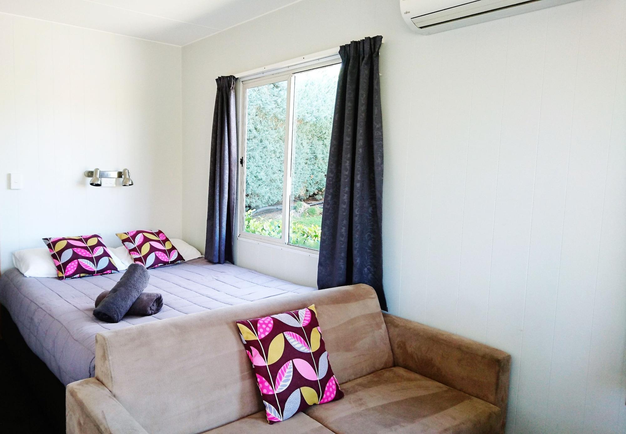 Taupo Debretts Spa Resort Luaran gambar The photo shows a cozy room with a bed and a small sofa. The bed features two decorative pillows with colorful patterns, and there is a rolled-up towel placed on the bed. Large windows are covered by dark curtains, allowing some natural light to ente