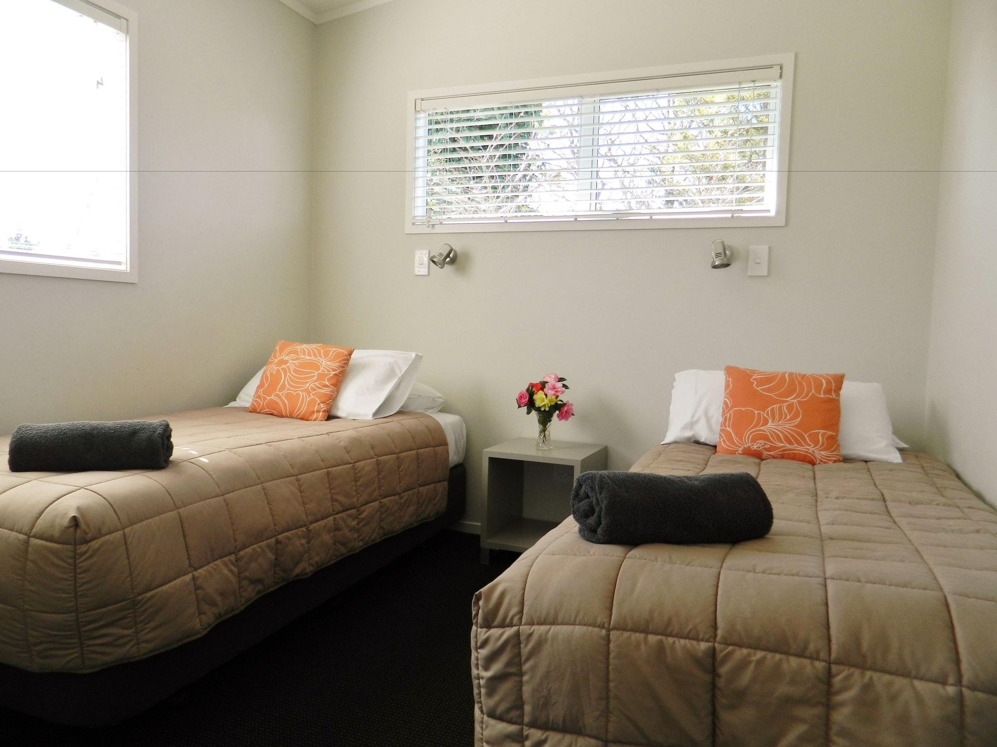 Taupo Debretts Spa Resort Luaran gambar The image shows a small, cozy room featuring two single beds. Each bed is dressed with a beige quilt and has an orange decorative pillow. There are two rolled towels placed on the beds. A small bedside table is located between the beds, with a vase o