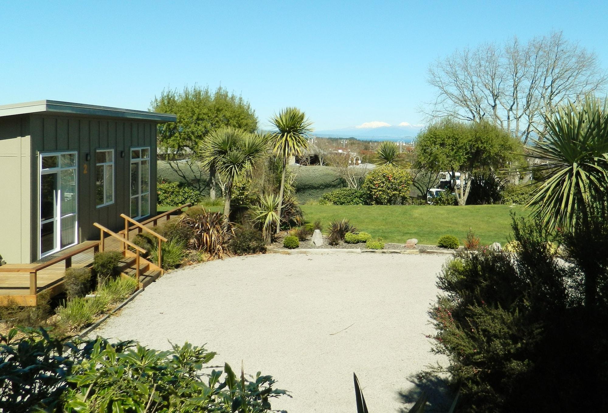 Taupo Debretts Spa Resort Luaran gambar The photo shows a scenic outdoor setting with a well-maintained garden area. In the foreground, there is a gravel patio or pathway surrounded by various plants and landscaping features. To the left, a small building with wooden steps leads up to it, 