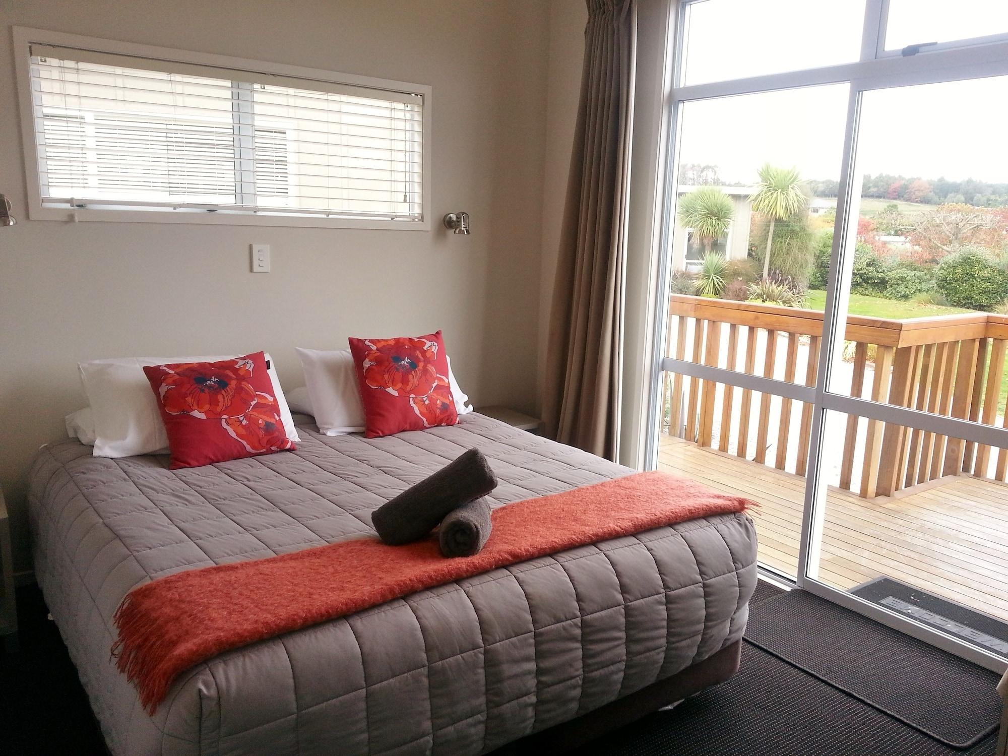 Taupo Debretts Spa Resort Luaran gambar The photo shows a cozy bedroom with a large bed centered in the room. The bed is covered with a light gray quilt and has decorative pillows in shades of red and orange. There is a bright orange throw blanket at the foot of the bed. A rolled towel is 