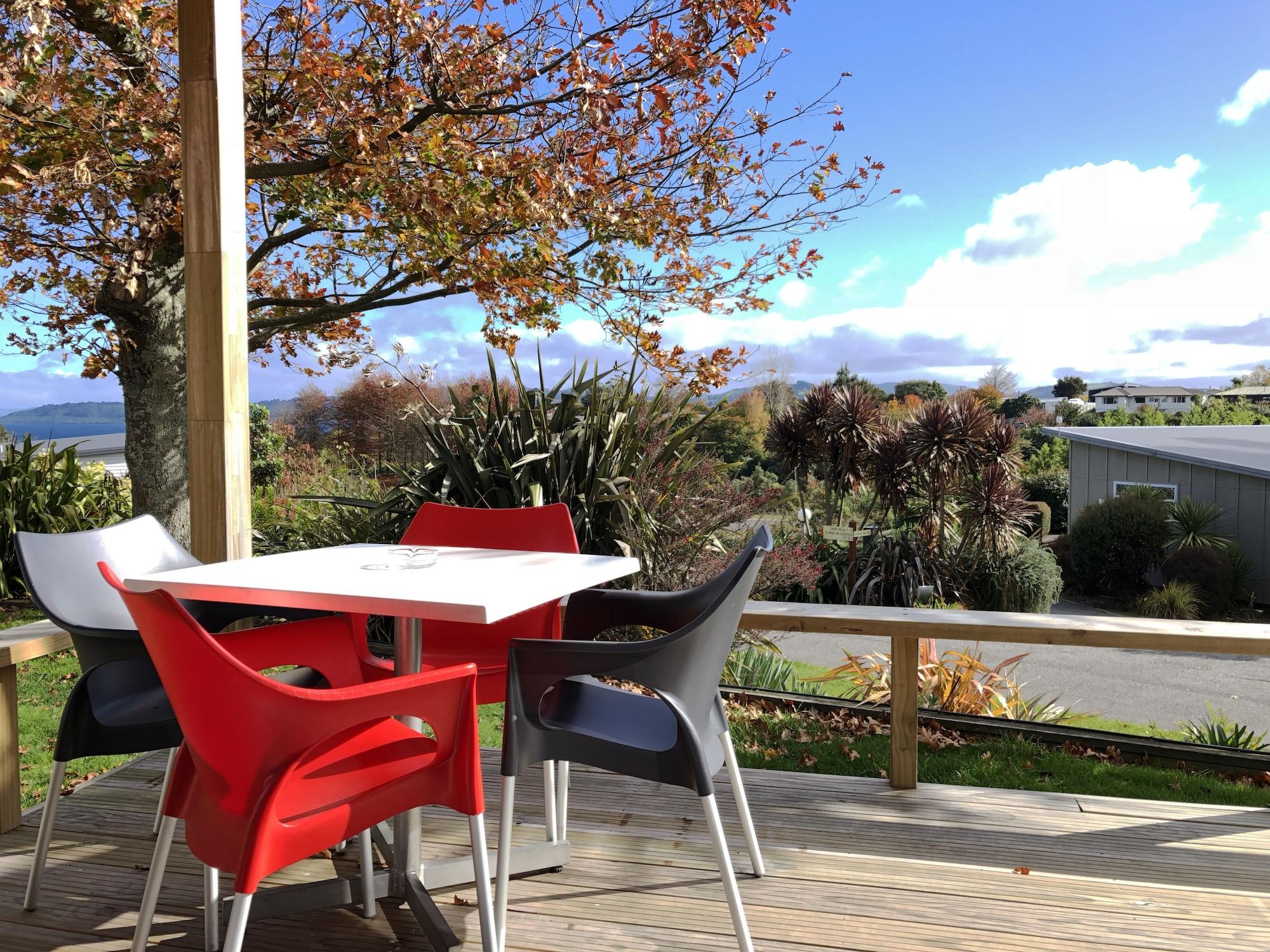 Taupo Debretts Spa Resort Luaran gambar The photo shows a picturesque outdoor seating area on a deck. There is a table surrounded by several chairs, with a couple of them in red and the others in gray and white. In the background, there are trees displaying autumn foliage, and beyond that,