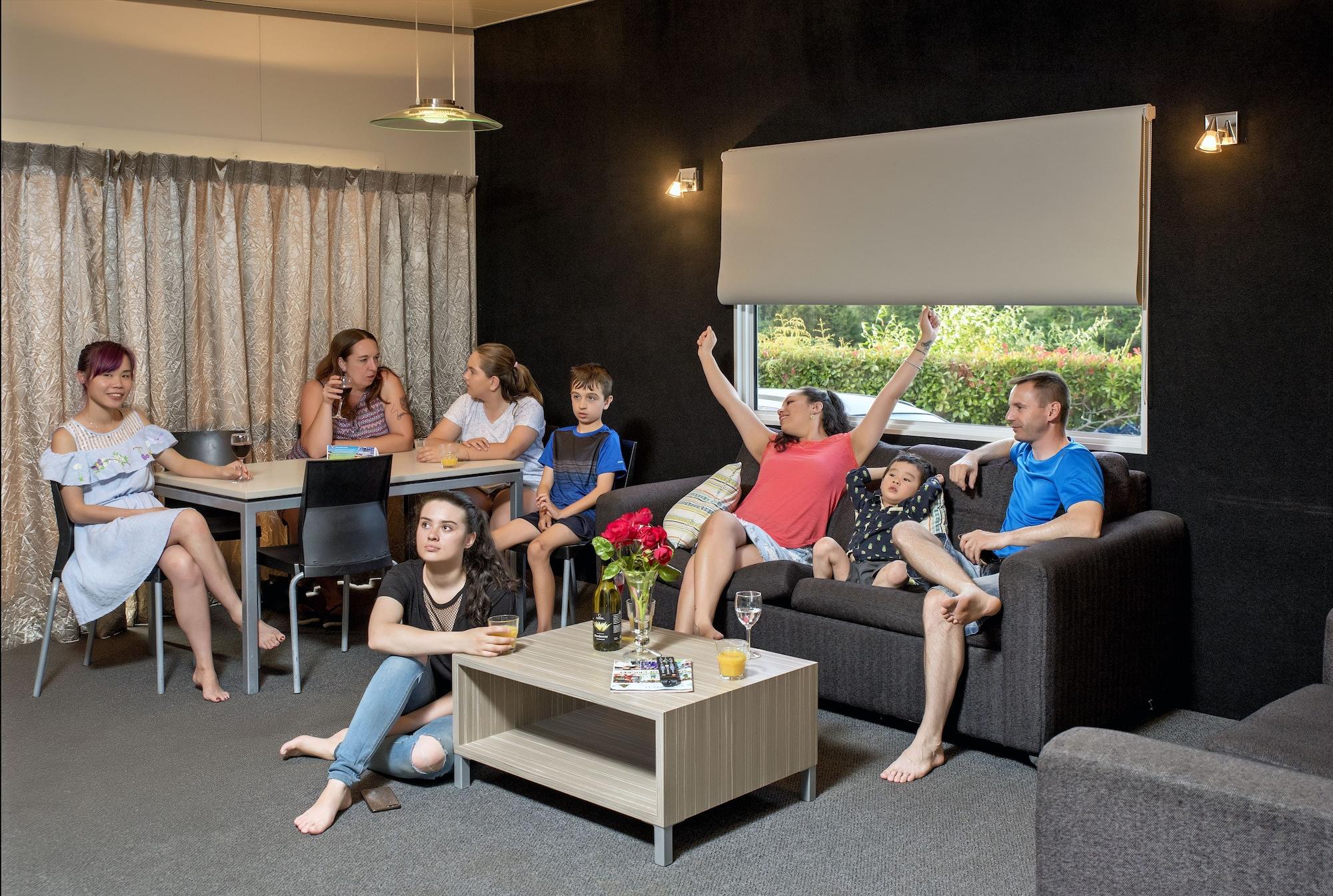 Taupo Debretts Spa Resort Luaran gambar The photo shows a group of people gathered in a cozy living room. There are several adults and children interacting with each other. One woman in a red top appears to be celebrating or expressing excitement, while others are engaged in conversation o