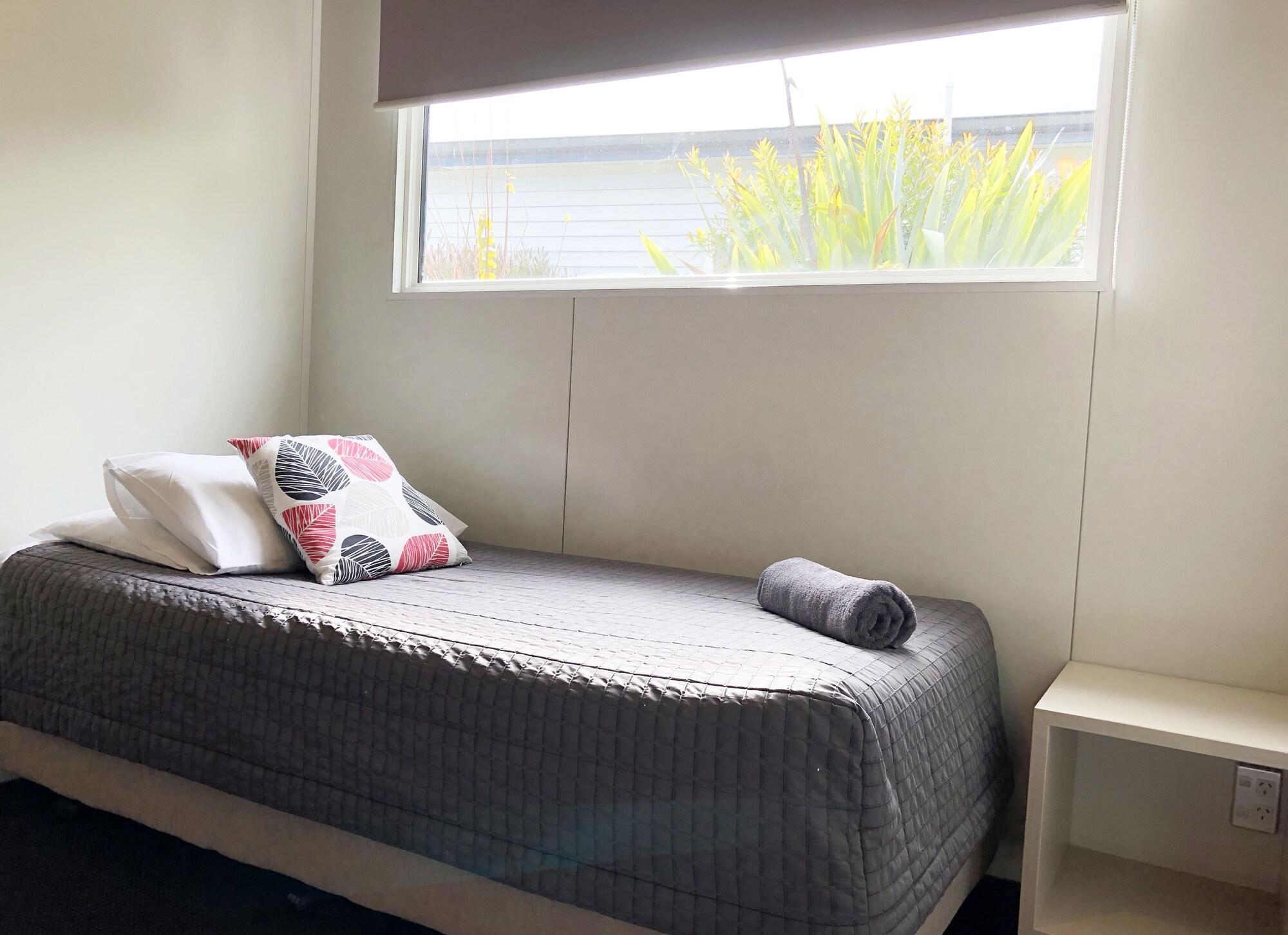 Taupo Debretts Spa Resort Luaran gambar The photo shows a simple bedroom. It features a single bed with a gray blanket, a couple of pillows (one patterned and one plain), and a rolled-up towel placed at the foot of the bed. Next to the bed is a small white nightstand. A large window lets i