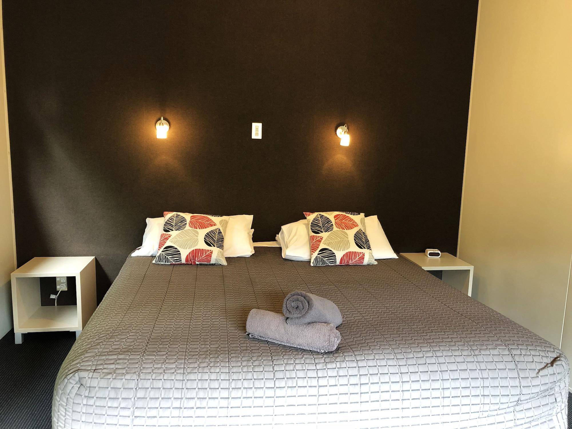 Taupo Debretts Spa Resort Luaran gambar The photo shows a bedroom with a large bed that has a gray quilted cover and decorative pillows. The walls behind the bed are painted a dark color, and there are two small wall-mounted lamps providing light. On either side of the bed, there are small