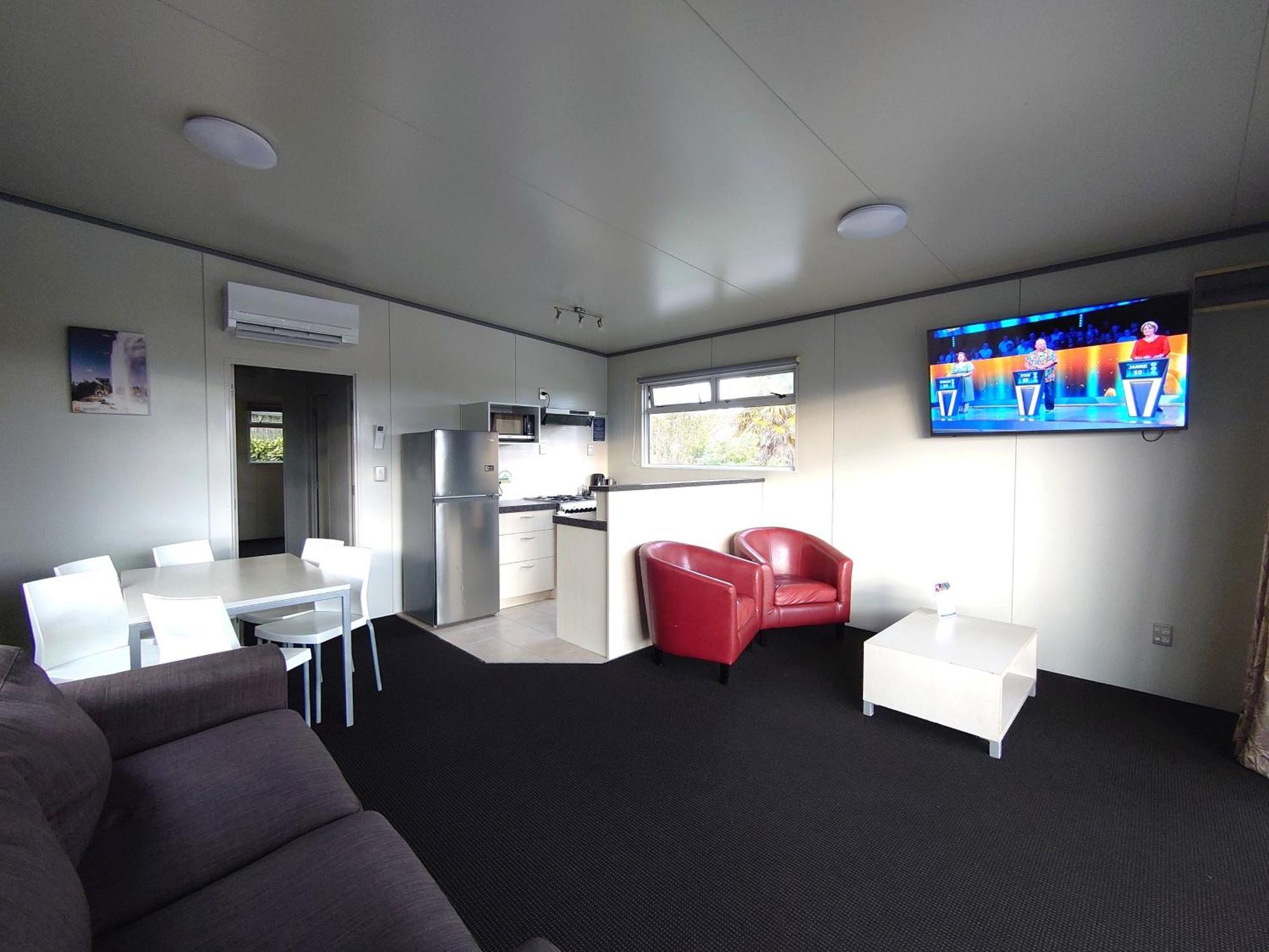 Taupo Debretts Spa Resort Luaran gambar The photo displays a modern, compact living space. It features a small kitchen area with a refrigerator and cabinets on one side. A dining table with white chairs is set up in the center of the room. On the opposite side, there is a comfortable dark 