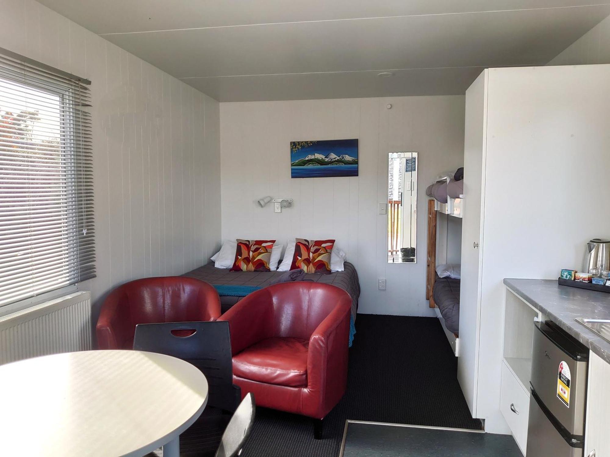 Taupo Debretts Spa Resort Luaran gambar The photo shows the interior of a small living space, possibly a cabin or studio apartment. There are two red armchairs and a round table in the foreground. The wall opposite has a bed with colorful pillows and a small nightstand. To the side, there 