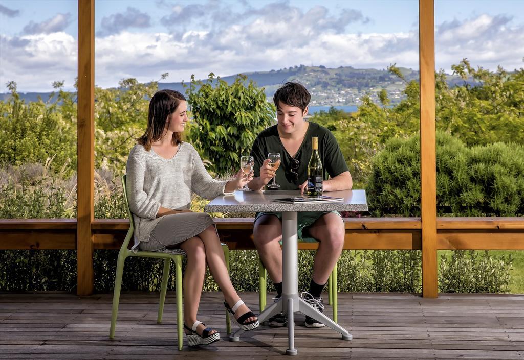 Taupo Debretts Spa Resort Luaran gambar The photo shows a young couple sitting at a table outdoors. They are enjoying drinks, with the woman wearing a light-colored sweater and a skirt, while the man is dressed in a dark shirt and shorts. The table is set with two glasses and a bottle of w