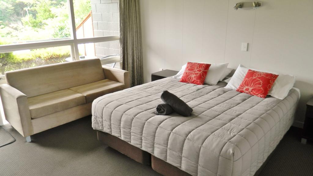 Taupo Debretts Spa Resort Luaran gambar The photo shows a cozy hotel or guest room. There is a large bed with gray bedding and two red decorative pillows. At the foot of the bed, there is a rolled-up towel. Next to the bed, there's a small bedside table and a light fixture on the wall. To 