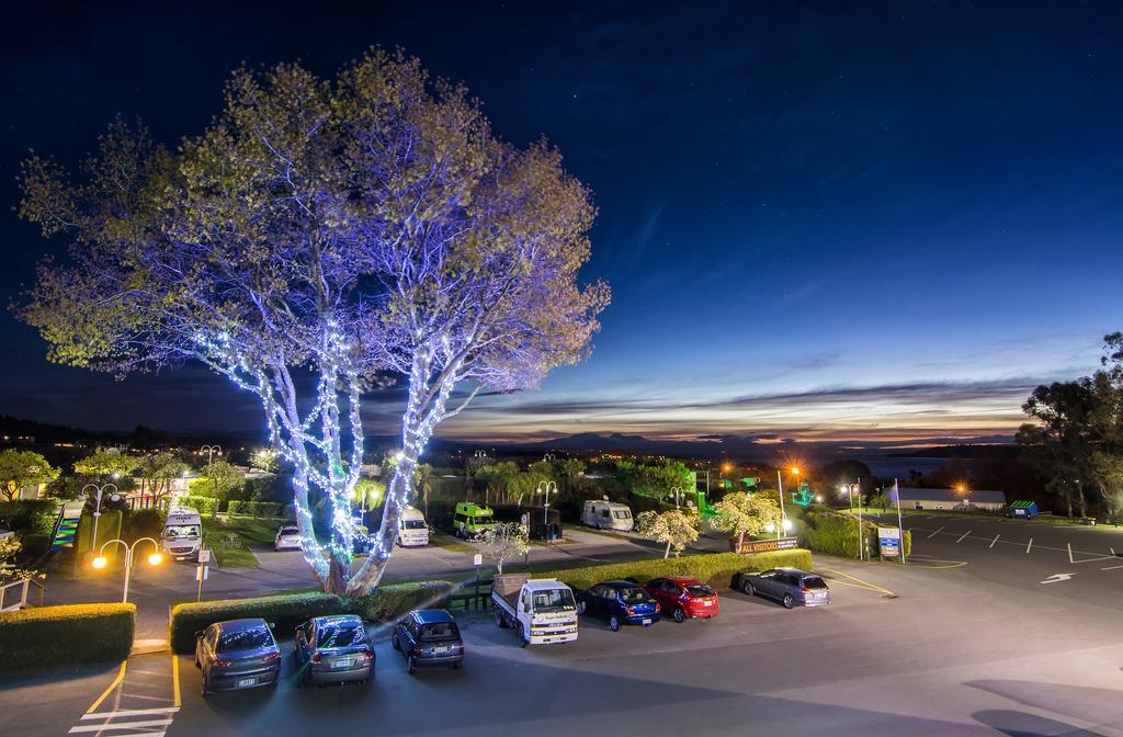 Taupo Debretts Spa Resort Luaran gambar The photo depicts a nighttime scene of a parking lot illuminated by streetlights. In the foreground, there are several parked cars in various colors. A large tree in the center is adorned with blue lights, creating a striking visual effect. The backg