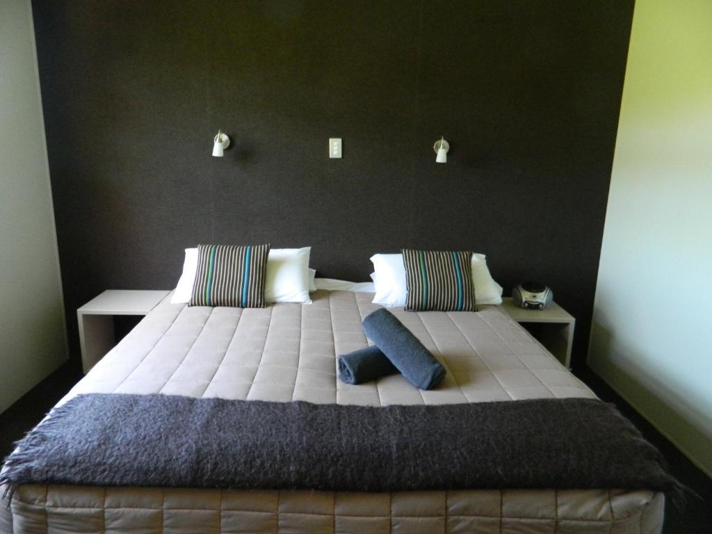 Taupo Debretts Spa Resort Luaran gambar The photo shows a modern bedroom featuring a large bed with a neutral-colored quilt and decorative pillows. There are two bedside tables on either side of the bed. The wall behind the bed is dark, creating a contrast with the lighter bedding. Additio